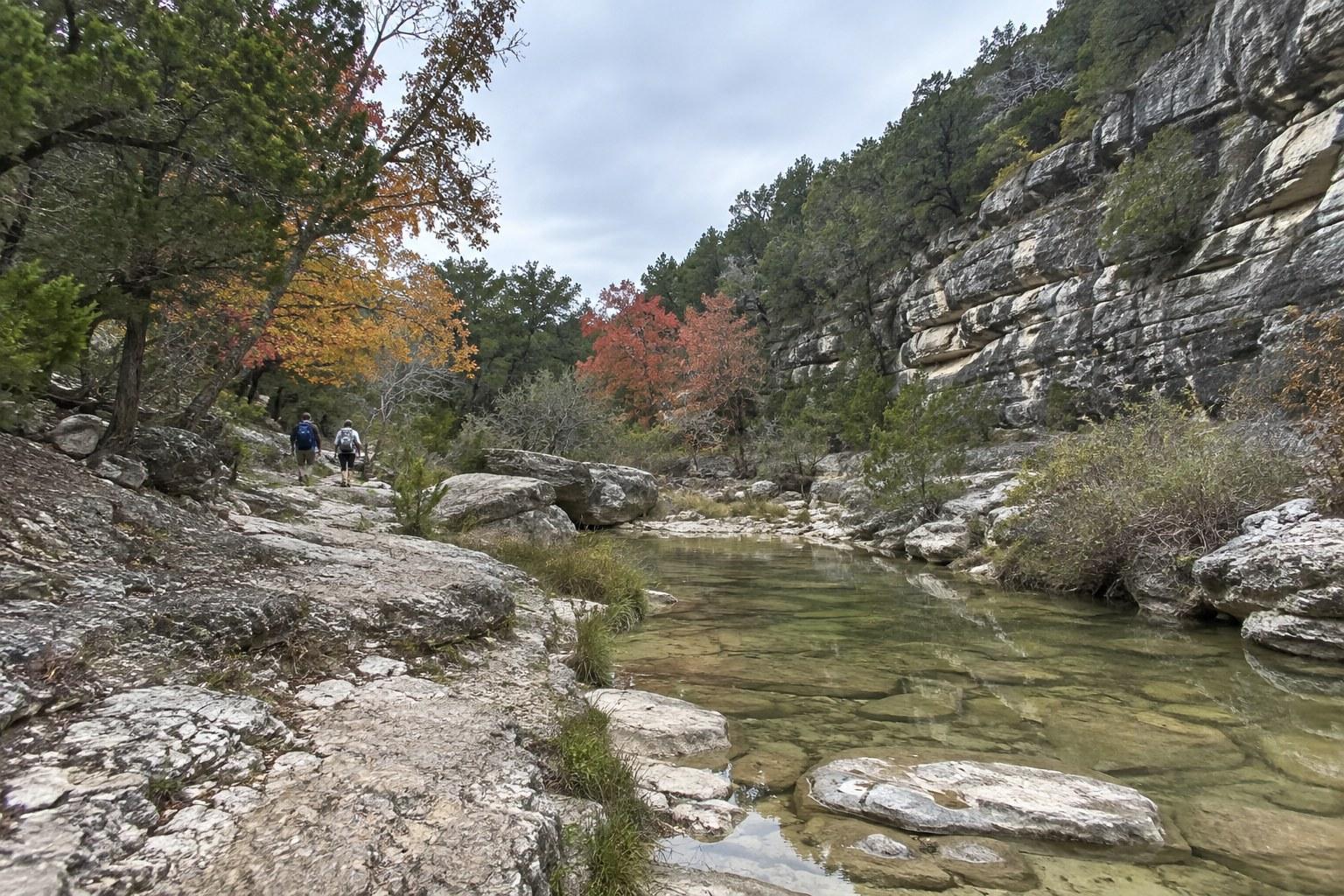 Hikers on a limestone trail beside a clear creek with fall color at Lost Maples in the Texas Hill Country